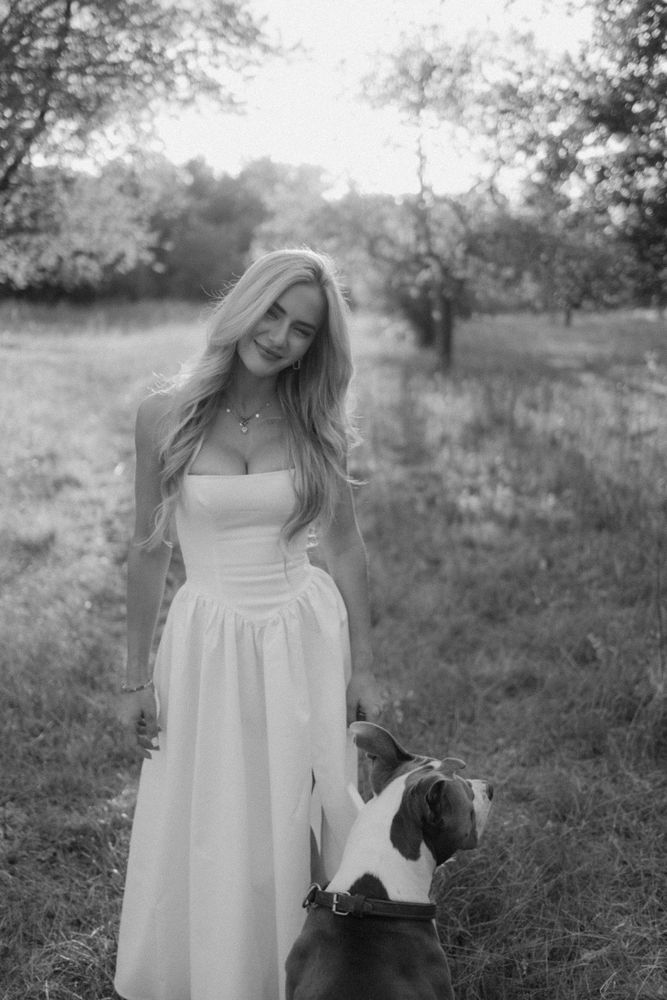 Stella im weißen Kleid auf einer Wiese, ihr Hund Tacco steht neben ihr